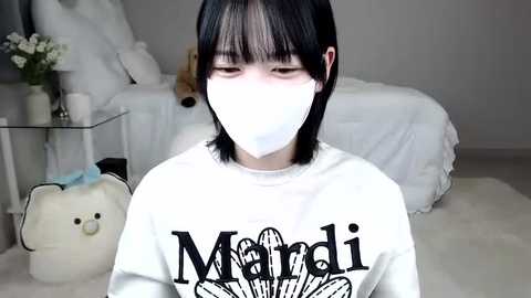 Media: Video of a young person with short black hair, wearing a white mask, a \"Mandy\" shirt, and sitting on a fluffy white rug in a minimalistic bedroom with a bed, plush bear, and flowers.