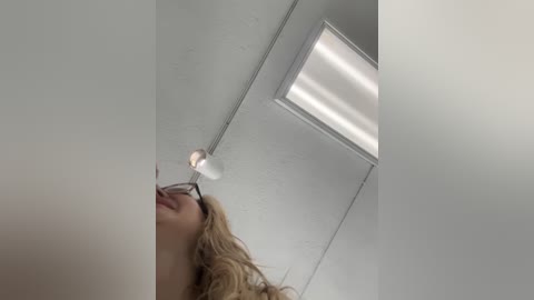 Media: A video of a blonde woman with glasses and light skin, smiling from the neck up, taken from a low angle. The background features a white ceiling with a rectangular fluorescent light and white walls.