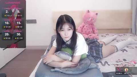 Media: Video of an Asian girl with long black hair, lying on a bed in a plaid skirt, holding a stuffed pink bear, in a bedroom with a digital clock and a stuffed animal.