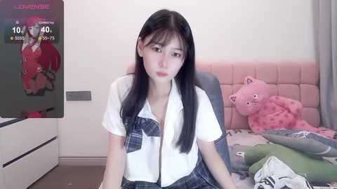 Media: A video of an East Asian woman with long black hair and fair skin, dressed in a white blouse and plaid skirt, sitting on a bed with pink cushions and a teddy bear, in a cozy bedroom.