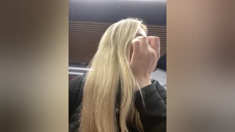 Media: Video of a blonde woman with long hair, wearing a black top, covering her face with both hands. She is seated indoors with a blurred background featuring a dark wooden surface and metallic elements.