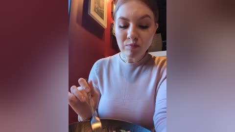 Media: A video of a young woman with light skin and brown hair, wearing a light pink long-sleeved shirt, eating with a spoon in a warmly lit room with red walls.