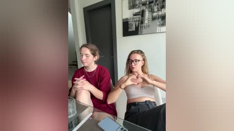 Media: Video of two young women in a modern living room, one with brown hair and red shirt, the other with blonde hair and glasses, forming heart shapes with their hands.