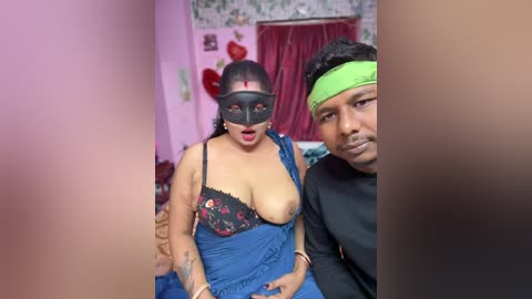 Media: Video of a South Asian woman with dark skin and medium build, wearing a black mask, blue dress, and black lace bra, sitting next to a man with medium skin and short black hair. The room has pink walls, a green headband, and a red curtain.