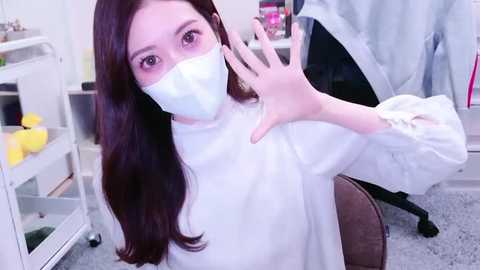 Media: A video of an Asian woman with long, dark hair, wearing a white lab coat, surgical mask, and gloves, making a peace sign in a laboratory setting with white walls and equipment.