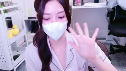 Media: Video of an Asian woman with long dark hair, wearing a white shirt and face mask, waving in a modern office setting.