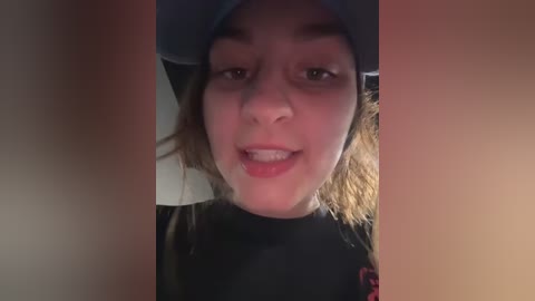 Media: Video of a young Caucasian woman with light skin, brown hair, and a blue baseball cap, smiling, wearing a black shirt, in a dimly lit room.