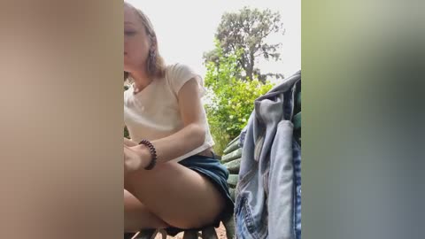 Media: Video of a young woman with fair skin, light brown hair, and a white t-shirt, sitting outdoors in a green, leafy environment, wearing denim shorts.