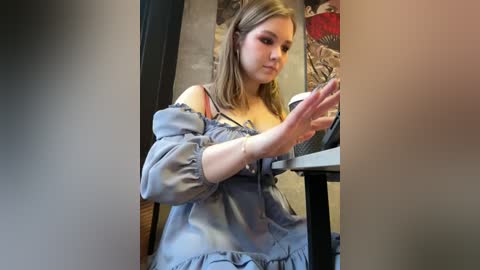 Media: Video of a young, fair-skinned woman with straight, shoulder-length blonde hair, wearing an off-shoulder, light blue blouse, holding a coffee cup. Background features a colorful, abstract painting and a wooden chair.