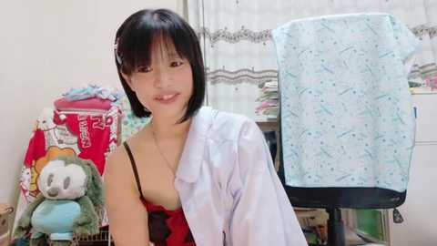 Media: A video of an East Asian woman with straight black hair, wearing a white lab coat over a red dress, standing in a cluttered bedroom with stuffed animals and a patterned shirt on a chair.