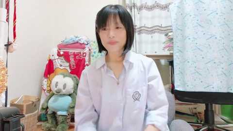 Media: A video of an Asian woman with short black hair in a white lab coat, sitting in a cluttered room with stuffed toys, a computer monitor, and a patterned cloth on a chair.