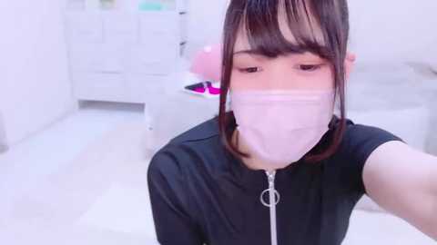 Media: Video of an East Asian woman with fair skin, wearing a black top and a pink face mask, in a bright, white room with minimalistic decor.