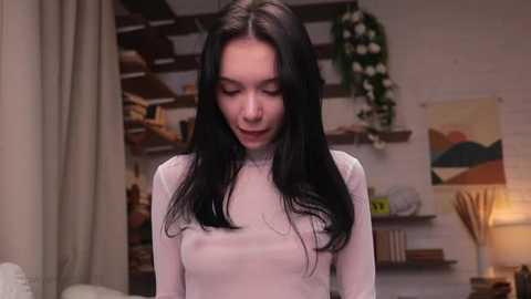 Media: Video of a young woman with long black hair, wearing a white long-sleeve top, standing in a cozy room with bookshelves, plants, and a painting.