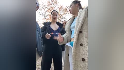 Media: Video of two women in a park. One woman in black hoodie and pants holds a phone, while the other in a beige coat and white top looks at her. Background features autumn trees and blue sky.