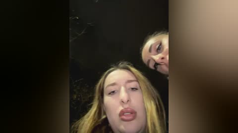 Media: Video of two young women with light skin and blonde hair, both with full lips, partially obscured by a dark, blurry background.