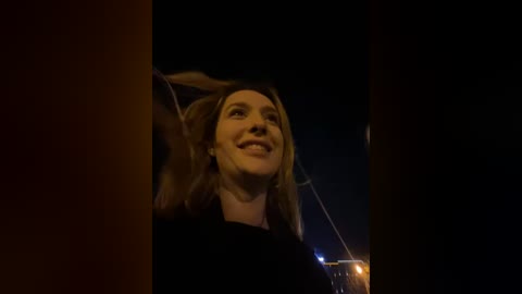 Media: Video of a smiling woman with long blonde hair, standing outdoors at night, dimly lit by a nearby light source.