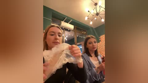 Media: Video of two women eating pizza in a cozy, warmly lit restaurant with brick walls and chandelier. One woman has blonde hair, the other brunette.