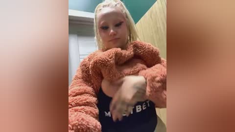 Media: Video of a blonde girl with braids wearing a fluffy, orange sweater, holding a stuffed animal. The background features green and beige walls.