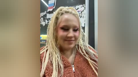 Media: Video of a smiling, light-skinned woman with blonde dreadlocks, wearing a rust-colored sweater. Background features a black wall with white and red graffiti.