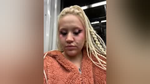 Media: Video of a young blonde woman with braided hair, wearing an orange fuzzy robe, crying with mascara running down her face, in a dimly lit, dark subway car.