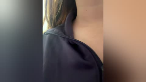 Media: Video of a woman's bare shoulder and chest in a black zip-up hoodie, revealing light skin and a hint of cleavage, blurred background.