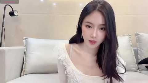 Media: Video of an East Asian woman with long black hair, fair skin, and light makeup, wearing a white off-shoulder lace top, seated on a light grey sofa with decorative pillows. The background features a modern, minimalist room with a black floor lamp.