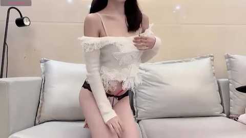 Media: Video of an East Asian woman with fair skin and straight black hair, wearing a white off-shoulder lace top and black lace panties, sitting on a light gray couch with soft pillows.