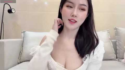 Media: Video of an Asian woman with fair skin and long black hair, wearing a white lace bra and a white sweater, sitting on a beige couch, touching her cheek, in a softly lit room.