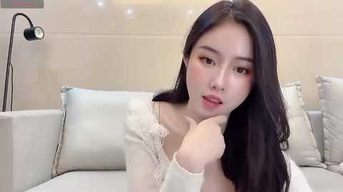 Media: A video of an East Asian woman with long black hair, wearing a white off-shoulder lace top, sitting on a light grey couch, with a black floor lamp in the background.