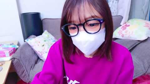 Media: Video of a woman with shoulder-length brown hair, wearing black-framed glasses, white face mask, and a bright pink sweater, sitting on a grey sofa with colorful pillows in a brightly lit room.