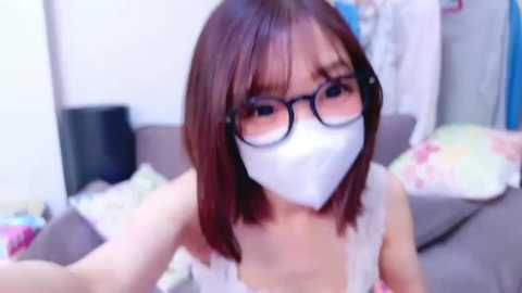 Media: A video of a young Asian woman with shoulder-length brown hair, wearing glasses and a white face mask, sitting on a bed with colorful pillows in a cozy, cluttered bedroom.