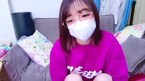 Media: Video of an Asian woman with glasses, wearing a pink sweatshirt, white mask, and pink pants, sitting on a gray couch, surrounded by colorful pillows in a cluttered room.