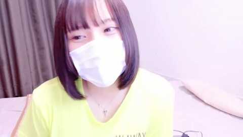 Media: A video of an East Asian woman with straight black hair and bangs, wearing a yellow T-shirt and white face mask, sitting indoors with beige curtains in the background.