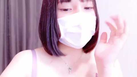 Media: Video of an East Asian woman with a bob haircut, wearing a white face mask and a lavender tank top. She is indoors with a blurred background, raising her hand.