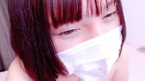 Media: Video of a young Asian woman with straight black hair and bangs, wearing a white surgical mask, looking tired, in a blurred indoor setting.