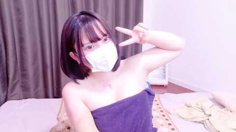 Media: Video of an Asian woman with straight black hair, wearing a face mask, purple towel, and giving a peace sign, indoors, on a tatami mat.