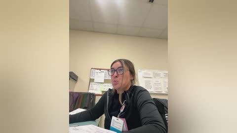 Media: A video of a woman with glasses and a badge, seated at a desk in an office, surrounded by paperwork and a bulletin board.