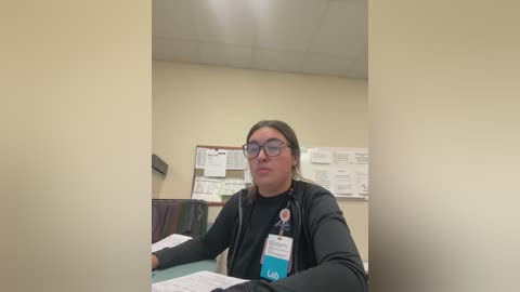 Media: Video of a middle-aged woman with glasses, dark hair, wearing a black zip-up jacket and a lanyard with a badge, seated at a desk in an office with beige walls and a bulletin board.