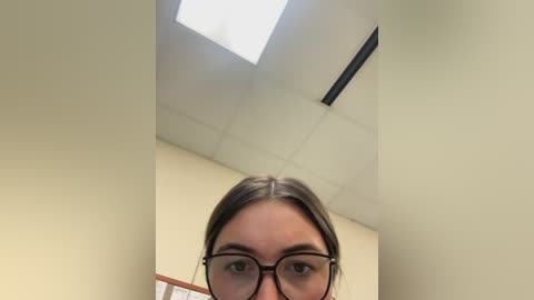 Media: Video of a young woman with light skin and brown hair, wearing black-framed glasses, standing in a fluorescent-lit hallway. The ceiling has a rectangular skylight, and beige walls surround her.