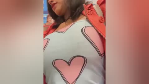 Media: Video of a woman with medium brown skin, wearing a white shirt with pink heart designs, red jacket, and dark hair, indoors with a blurred background.