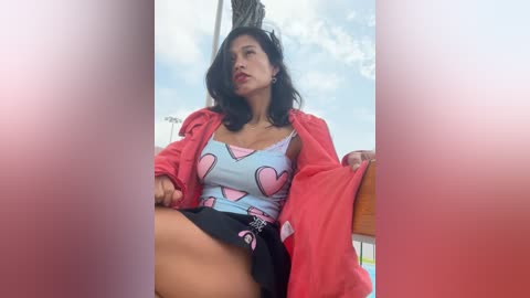 Media: Video of a woman with medium skin tone, wearing a red jacket over a pink and blue top with heart patterns, sitting on a boat deck.