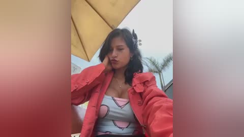 Media: Video of a young woman with medium skin tone, medium build, and medium-sized breasts, wearing a pink jacket over a pink heart-patterned top, sitting under a yellow umbrella, with blurred palm trees in the background.