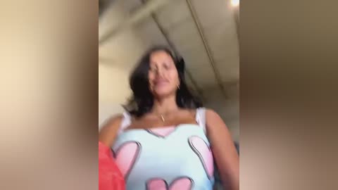 Media: Video of a woman with long black hair, wearing a light blue top with pink heart patterns, standing in a room with beige walls and a white ceiling.