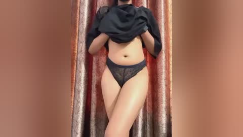 Media: A video of a woman lifting a black shirt to reveal her bare midriff and wearing dark blue lace panties, standing in front of a draped curtain with a gradient of pink and brown.