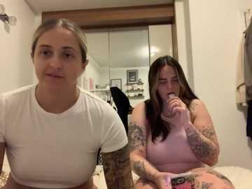 Media: Video of two women in a bathroom: one with light skin and brown hair in a white crop top, the other with dark hair and tattoos, wearing a pink tank top, holding a phone.