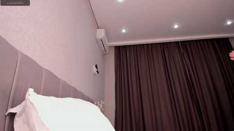 Media: Video of a modern, minimalist bedroom with white walls, a white headboard, and dark brown curtains. An air conditioning unit is mounted on the wall, and recessed ceiling lights provide soft illumination.