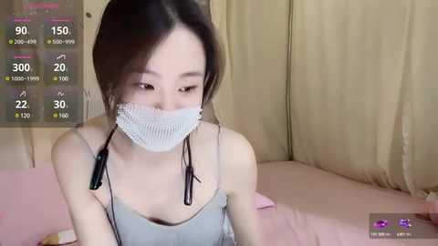 Media: Video of an Asian woman with light skin and long dark hair, wearing a white medical mask and a gray spaghetti-strap tank top. She's indoors, sitting on a pink bed, with a digital health monitoring app screen visible.