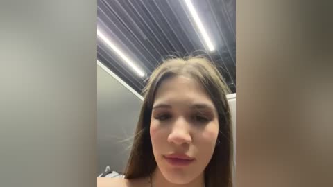 Media: Video of a young woman with long, straight brown hair, fair skin, and subtle makeup, standing in a modern, dimly lit room with a metallic ceiling.