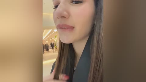 Media: Video of a young woman with long, straight brown hair, fair skin, and full lips, wearing a dark top. She stands in a modern, brightly lit shopping mall, blurred background with shoppers and stores.