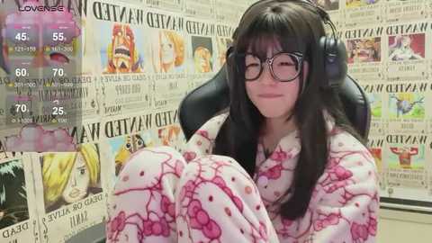 Media: Video of a smiling woman with long black hair, wearing glasses and a Hello Kitty pajama set, sitting on a black chair in a room filled with newspaper clippings.